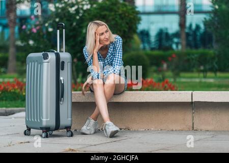 Triste giovane donna bionda in pantaloncini, una camicia è seduta con una valigia su una panca contro lo sfondo di alberi, cespugli in una giornata estiva Foto Stock