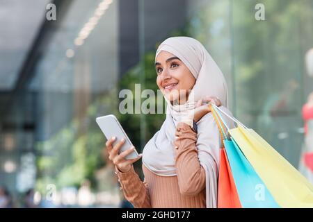 Sorridente signora araba millennial graziosa in hijab gode dello shopping in linea con i sacchetti, surf sul telefono Foto Stock