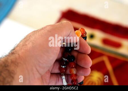 Una mano tiene un rosario islamico colorato su uno sfondo sfocato di un tappeto di preghiera in una moschea, le perle di preghiera islamica chiamato Misbaha o Tasbih, preghiera Foto Stock