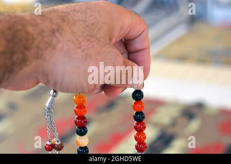 Una mano tiene un rosario islamico colorato su uno sfondo sfocato di un tappeto di preghiera in una moschea, le perle di preghiera islamica chiamato Misbaha o Tasbih, preghiera Foto Stock