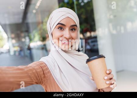 Sorridente bellezza giovane arabo islamico signora in hijab con un sacco di borse rende selfie sul dispositivo vicino al centro commerciale Foto Stock