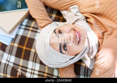 Soddisfatto felice giovane araba musulmana donna in hijab riposo nel parco, si trova in plaid nel giardino universitario Foto Stock