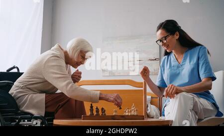 donna anziana felice e infermiera sorridente che gioca a scacchi in casa di cura Foto Stock