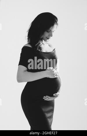 Bella giovane donna incinta che abbraccia il ventre in posa in studio, foto in bianco e nero. Buona gravidanza e maternità Foto Stock