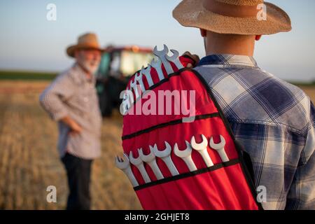 Vista posteriore del giovane agricoltore che tiene un set di chiavi sulla spalla e che cammina verso il trattore rotto sul campo Foto Stock