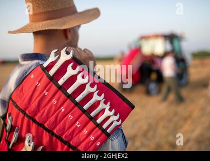 Giovane agricoltore che tiene un set di chiavi sulla spalla e cammina verso il trattore rotto sul campo Foto Stock