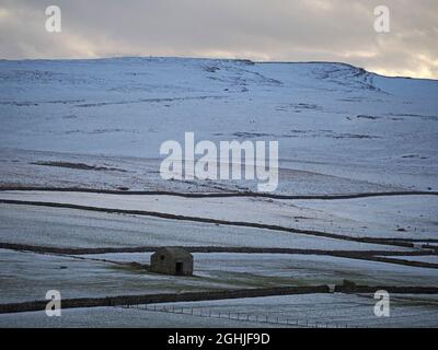 Paesaggio invernale con fienile in campi innevati fiancheggiati da recinzioni e muri sotto le campane lontane dell'altopiano punteggiate da piccole figure a piedi, Cumbria, Inghilterra, Regno Unito Foto Stock