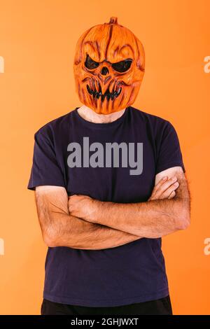 Uomo che indossa una maschera in lattice di zucca spaventosa con t-shirt blu a braccia incrociate, su sfondo arancione. Halloween e giorni del concetto morto. Foto Stock