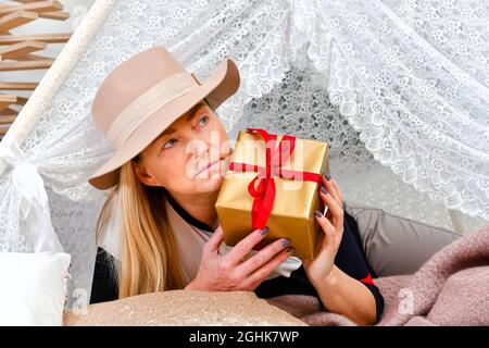 Donna di mezza età con cappello elegante che posa tra le scatole regalo di natale in una stanza decorata, concetto di Natale Foto Stock