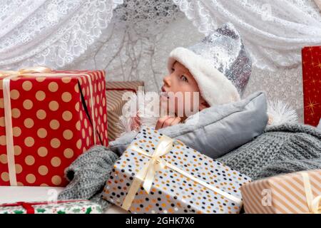 Ritratto di un bambino carino bambina che indossa un cappello di Natale color argento che giace tra le scatole del regalo, concetto di Natale Foto Stock