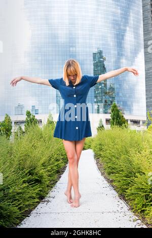 Corpo pieno di giovane donna a piedi nudi in abito blu che balla su punta di lupo con le braccia distese su uno stretto sentiero contro l'edificio urbano contemporaneo con g Foto Stock