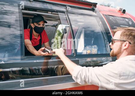 Giovane uomo che tiene lo smartphone sul terminale di pagamento mentre paga il fast food Foto Stock