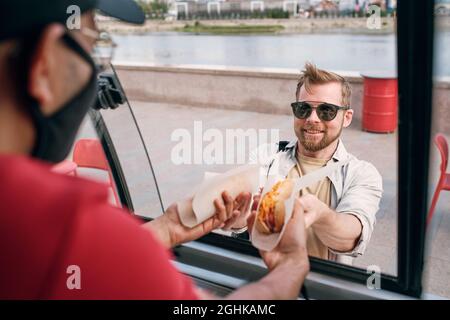 Felice uomo giovane in occhiali da sole che acquista due hamburger in camion Street food Foto Stock