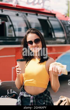 Felice giovane donna brunette in occhiali da sole che tengono hamburger e bevanda acquistati in strada cibo camion Foto Stock