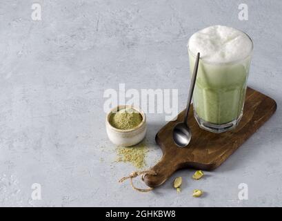 Bevete il caffè latte Matcha in un bicchiere di vetro su un asse di legno con un cucchiaino su sfondo di cemento. Il concetto di bevande vegetariane sane Foto Stock