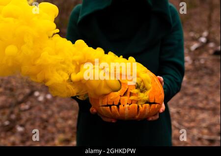 Una strega creepy tiene una zucca fumante in una foresta profonda. Jack o lanterna emette fumo giallo per Halloween Foto Stock