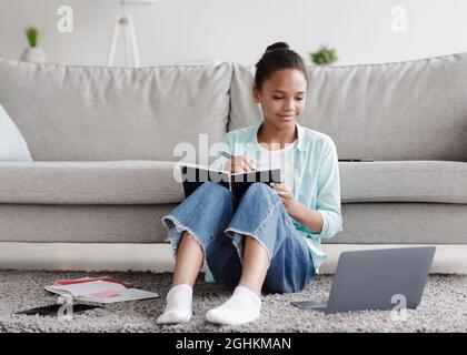 Sorridente adolescente africano americano studia da remoto, scrive in notebook, si siede sul pavimento e guarda il portatile Foto Stock