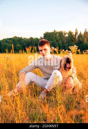 Un ragazzo di 18 anni è seduto con un cane su un campo in erba fitta in una serata estiva. Foto Stock