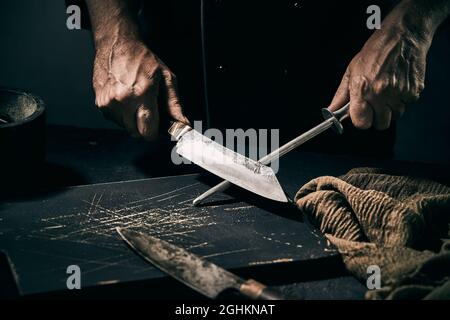 Lo chef affila la lama di un grande coltello su una lima metallica portatile su un tagliere in luce oscura e oscura con copyspace Foto Stock