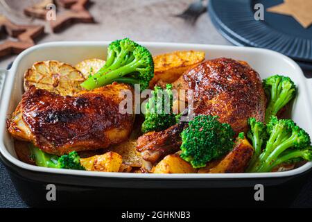 Cosce di pollo al forno con patate, broccoli e aglio in piatto da forno, sfondo scuro natale. Sano concetto di cibo festivo. Foto Stock