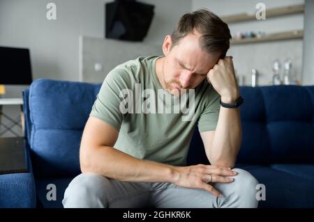 Il giovane uomo depresso che tiene la testa in mani seduto sul divano a casa da solo, ha problemi, soffrendo di forte mal di testa o di rammarico per errore, sconvolto ragazzo solitario preoccupante Foto Stock
