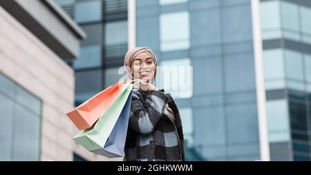 Felice attraente musulmano afro americano donna acquirente in hijab e cappotto con pacchetti con acquisti su sfondo negozio Foto Stock