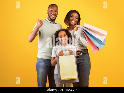 Concetto di black friday. I genitori neri felici e la figlia cute che tiene i sacchetti del compratore, l'uomo che mostra il pollice in su Foto Stock