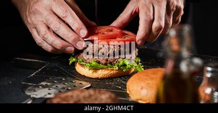 Lo chef prepara un doppio hamburger di manzo da asporto con polpettine di carne macinata e fette di pomodoro fresco in un primo piano sulle sue mani e sul cibo Foto Stock