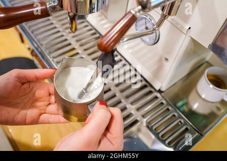 Barista femminile anonima con raccolto ad alto angolo che monta latte in caraffa metallica con macchina da caffè professionale mentre prepara bevande in una moderna caffetteria Foto Stock