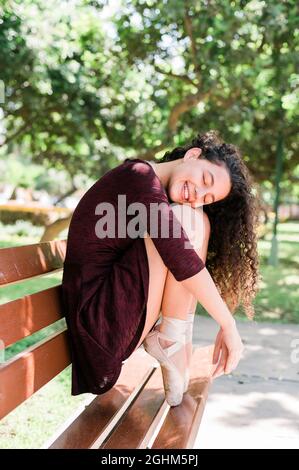 Giovane bella ballerina in abito nero seduta su una panca in un parco con gli occhi chiusi e sorridenti Foto Stock