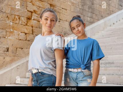 Madre e figlia in piedi su scale di pietra fuori. Una donna di mezza età e una bambina che si tengono le mani e indossano magliette blu e guardando Foto Stock