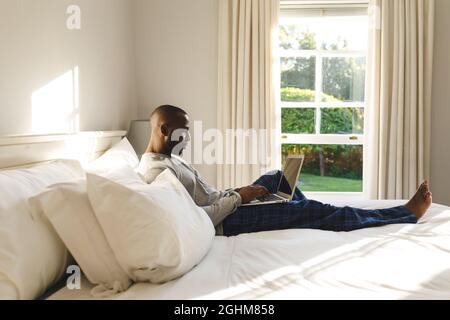 Uomo afroamericano che usa un computer portatile e che si trova a letto nella sua camera da letto Foto Stock