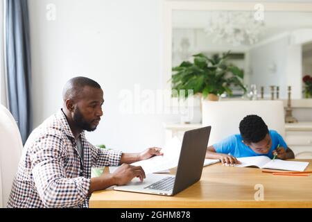 Il padre afroamericano che lavora sul computer portatile in sala da pranzo con il figlio seduto con lui che fa i compiti Foto Stock