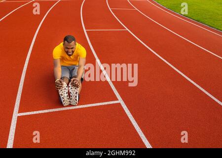 Atletica maschile in giallo abbigliamento sportivo seduto sul tracciato dello stadio, facendo un riscaldamento prima di correre, allungando le gambe. Uomo africano americano concentrato Foto Stock