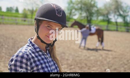 una giovane ragazza con un casco sulla testa guarda una ragazza sfocata in piedi accanto ad un cavallo marrone e guarda la macchina fotografica. Foto di alta qualità Foto Stock