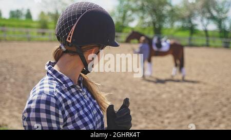 una giovane ragazza con un casco sulla testa guarda una ragazza sfocata in piedi accanto ad un cavallo marrone. Foto di alta qualità Foto Stock