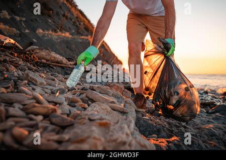 Giornata mondiale dell'ambiente. Un attivista maschile prende una bottiglia di plastica sulla spiaggia. Sullo sfondo, il mare e il tramonto. Vista ad angolo basso. Il concetto Foto Stock