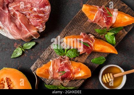 Prosciutto con fette di melone cantalupo, miele e basilico sul tagliere su sfondo scuro. Antipasto italiano. Vista dall'alto. Cibo sano Foto Stock