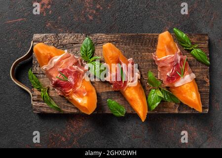 Fette di melone cantalupo con prosciutto e basilico su sfondo nero. Antipasto italiano. Vista dall'alto. Cibo sano Foto Stock