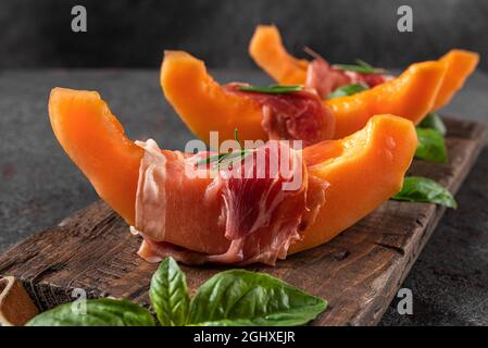 Melone cantalupo fresco con prosciutto e basilico su sfondo nero. Antipasto di cucina italiana. Primo piano Foto Stock