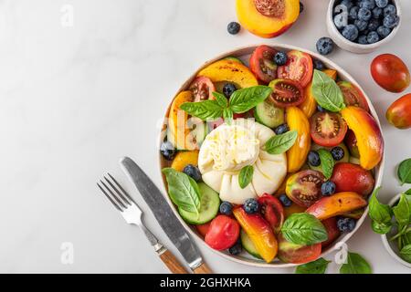 Insalata di burrata con pesche alla griglia, pomodori, mirtilli, cetrioli e basilico in un piatto con posate su tavola bianca. Vista dall'alto con spazio di copia. Foto Stock