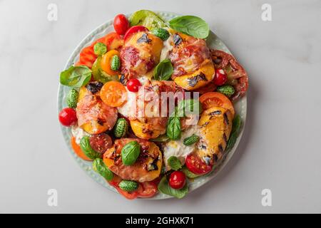 Pesche alla griglia e insalata di prosciutto con burrata, pomodori, cetrioli e basilico in un piatto su tavola bianca. Vista dall'alto. Dieta sana concetto di cibo Foto Stock