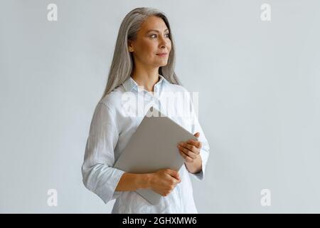 Positiva donna d'affari asiatica matura tiene il laptop in piedi su sfondo grigio chiaro Foto Stock