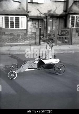 In questo quadro storico dei primi anni '60, un ragazzo adolescente che indossa un maglione a collo di tartaruga di lana seduto su un go-cart fatto in casa che corre lungo una strada suburbana, Inghilterra, Regno Unito. Traditonally fatto da pezzi vecchi di legno fissato a ruote di pam e sterzato con un po 'di corda, molti un giovane ha avuto grande divertimento con una tale macchina, che erano popolari alla fine degli anni '50/primi anni '60, quando erano comunemente noti come 'carrelli'. Foto Stock