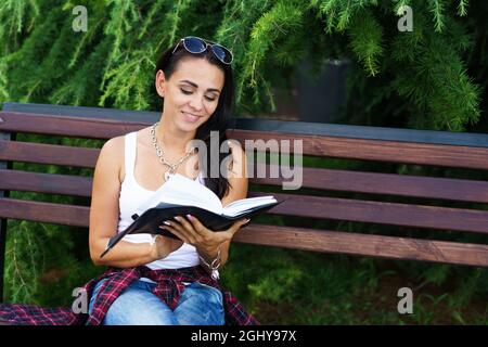 La ragazza caucasica freelance si siede sulla panca e scrive in un taccuino. Concetto di educazione e giornalismo. Donna felice e libera in abiti casual in parco sullo sfondo di cespugli verdi su panca di legno Foto Stock