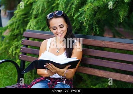 La ragazza caucasica freelance si siede sulla panca e scrive in un taccuino. Concetto di educazione e giornalismo. Donna felice e libera in abiti casual in parco sullo sfondo di cespugli verdi su panca di legno Foto Stock
