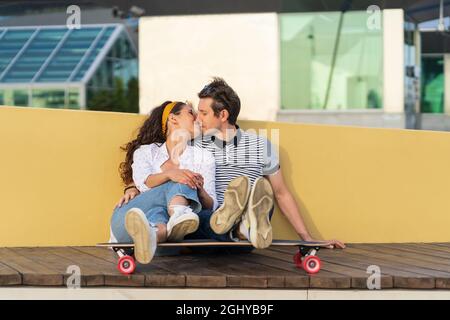 Un paio di pattinatori che si baciano si siedono a bordo lungo insieme all'aperto nello spazio urbano. Gioventù, romanticismo e amore Foto Stock