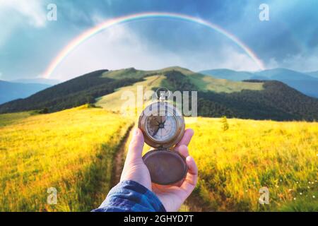 Uomo con vecchia bussola in metallo in mano su strada di montagna. Concetto di viaggio. Lussureggianti prati erbosi e arcobaleno sullo sfondo. Fotografia di paesaggio Foto Stock