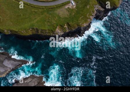 Vista panoramica della famosa scogliera di Moher in Irlanda con auto parcheggiata alla fine della strada sulla montagna con onde che colpiscono le rocce durante il giorno Foto Stock