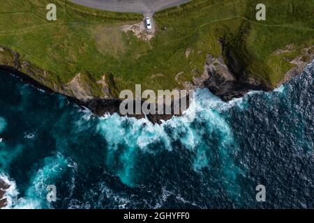 Vista panoramica dell'auto parcheggiata alla fine della famosa scogliera di Moher in Irlanda con onde che colpiscono le rocce durante il giorno Foto Stock